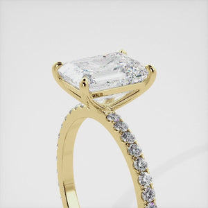 A close-up of an emerald solitaire lab-diamond ring with pave in yellow gold. The ring is set with an emerald-cut lab diamond in the center, surrounded by a row of smaller diamonds on the band. The diamond is climate-positive as it used carbon from the atmosphere to produce. The ring is available in a variety of sizes and starts at \\$6,350.00.
