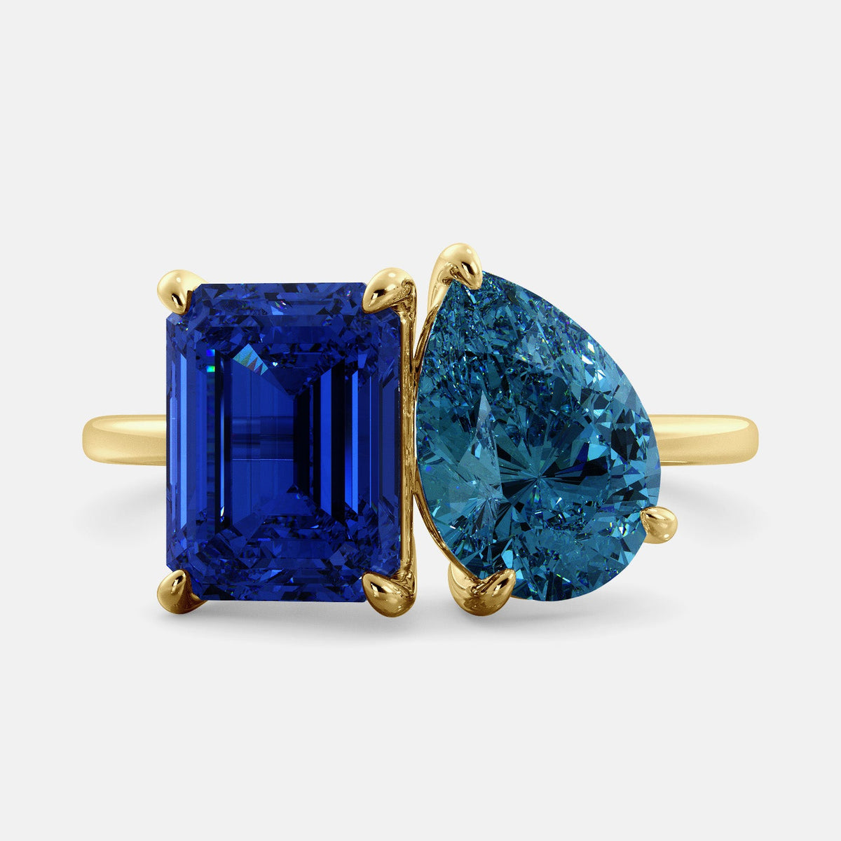 A toi et moi ring with an emerald-cut Blue Sapphire and pear-cut gemstone. The emerald-cut morganite is set on the left of the ring, and the pear-cut gemstone is set on the right. The ring is made of recycled yellow gold and has a simple, elegant design. The pear-cut gemstone can be customized. **Here are some additional details about the image:** * The emerald-cut white Blue Sapphire is a birthstone for September. * The pear-cut gemstone can be customized to any stone of your choice.