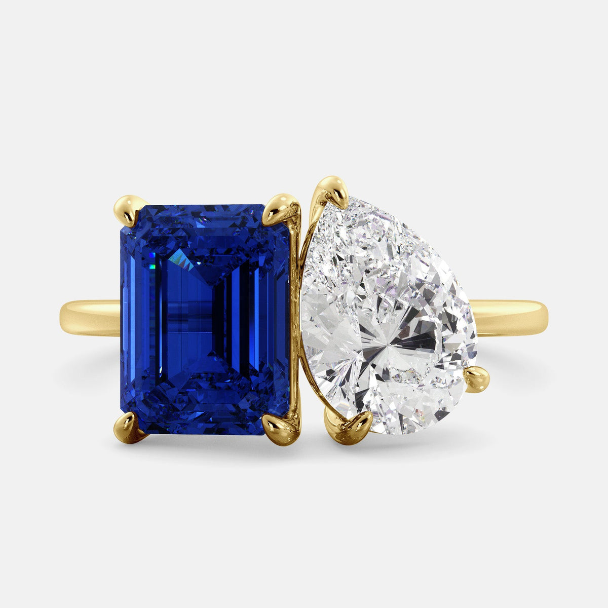 A toi et moi ring with an emerald-cut Blue Sapphire and pear-cut gemstone. The emerald-cut morganite is set on the left of the ring, and the pear-cut gemstone is set on the right. The ring is made of recycled yellow gold and has a simple, elegant design. The pear-cut gemstone can be customized. **Here are some additional details about the image:** * The emerald-cut white Blue Sapphire is a birthstone for September. * The pear-cut gemstone can be customized to any stone of your choice.