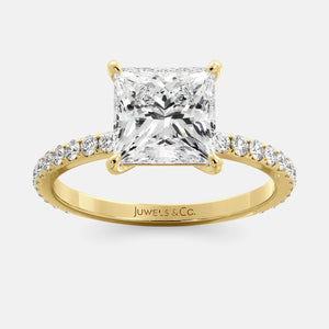 A close-up of a yellow gold cushion solitaire lab-grown diamond ring with an eternity band. The lab-grown diamond is a climate -positive diamond, created by using carbon from the atmosphere. The ring is set with a 1-carat cushion-cut diamond in the center, surrounded by a row of smaller diamonds on the band. The ring is available in a variety of sizes and starts at \\$6,400.00.