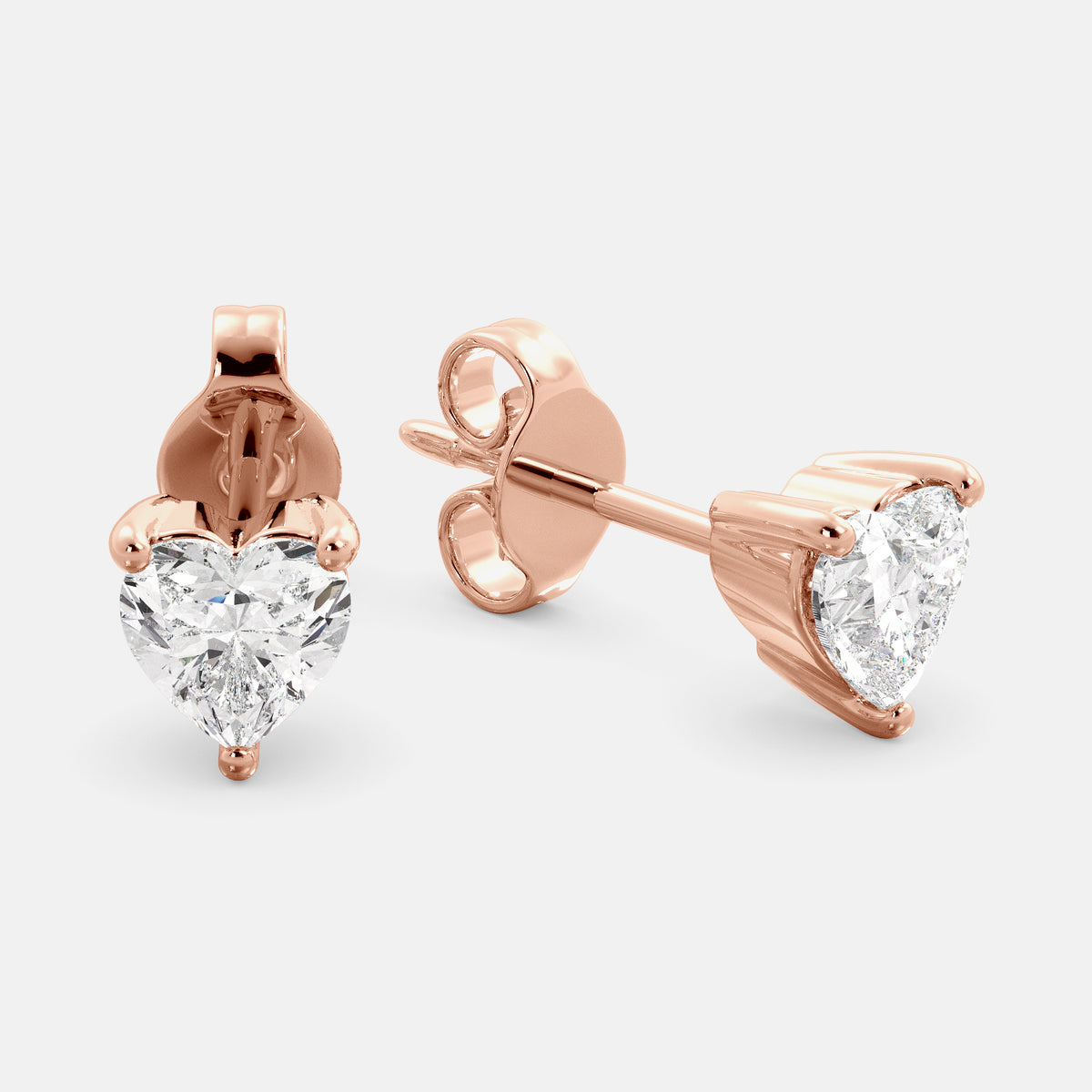A close-up of a pair of heart-shaped diamond stud earrings in recycled rose gold. Each earring is set with a single heart shape diamond in the center. The earrings are made of recycled rose gold and have a simple, elegant design. The diamonds are sparkling and reflect light beautifully. The earrings are a perfect gift for a loved one or a special occasion.