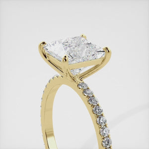 A close-up of a yellow gold cushion solitaire lab-grown diamond ring with an eternity band. The lab-grown diamond is a climate -positive diamond, created by using carbon from the atmosphere. The ring is set with a 1-carat cushion-cut diamond in the center, surrounded by a row of smaller diamonds on the band. The ring is available in a variety of sizes and starts at \\$6,400.00.