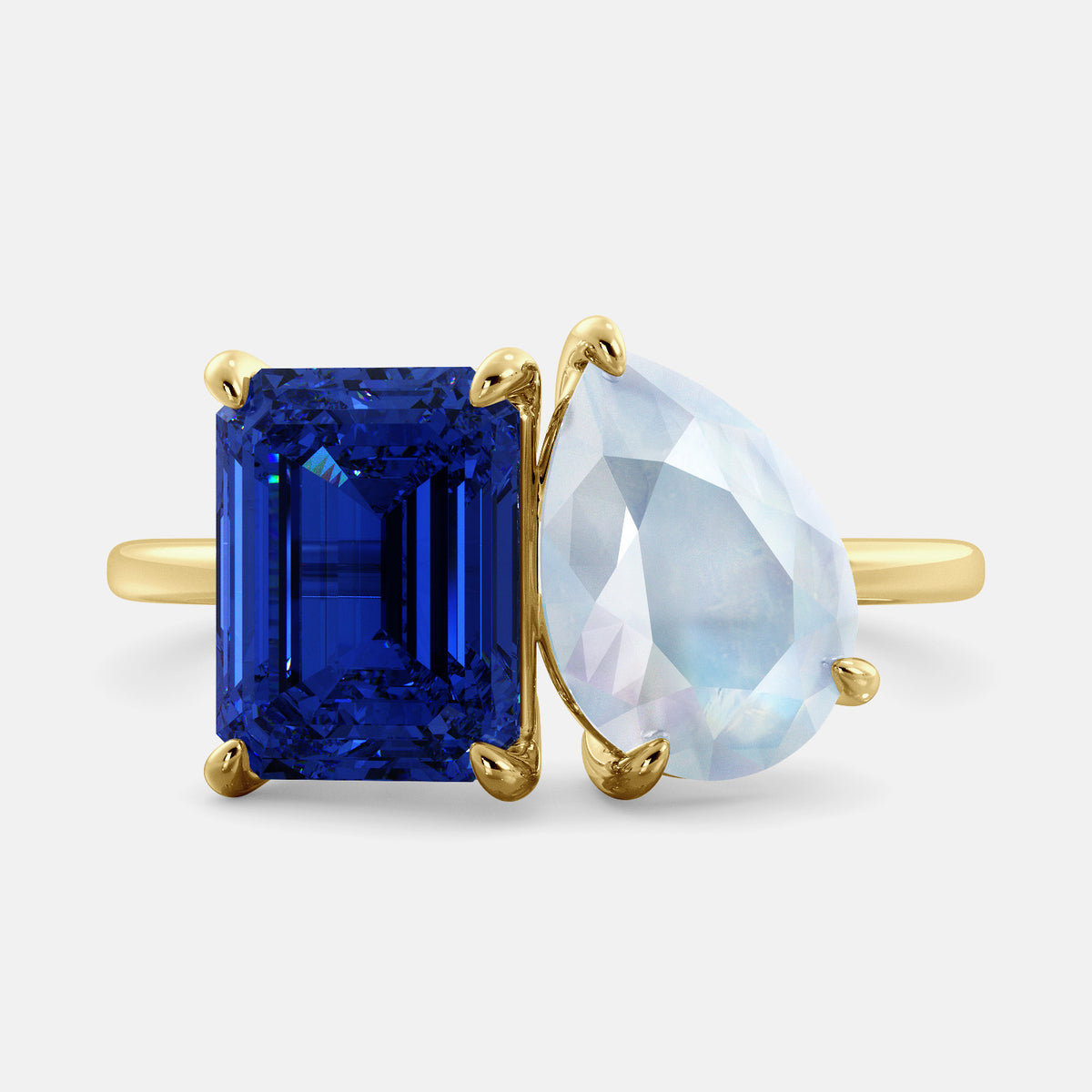 A toi et moi ring with an emerald-cut Blue Sapphire and pear-cut gemstone. The emerald-cut morganite is set on the left of the ring, and the pear-cut gemstone is set on the right. The ring is made of recycled yellow gold and has a simple, elegant design. The pear-cut gemstone can be customized. **Here are some additional details about the image:** * The emerald-cut white Blue Sapphire is a birthstone for September. * The pear-cut gemstone can be customized to any stone of your choice.