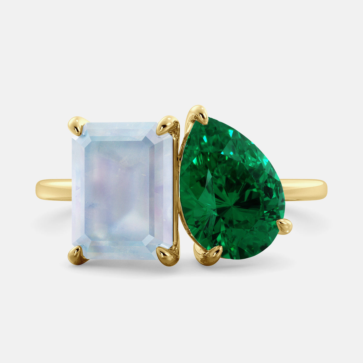 This image shows a toi et moi ring with a pear-shaped emerald and an emerald-cut moonstone. The ring is set in a 14k yellow gold band and is available in all sizes. The moonstone is a gemstone that is said to promote intuition, imagination, and creativity. The emerald is a gemstone that is said to promote love, compassion, and forgiveness. The toi et moi ring is a beautiful and unique way to show your love and commitment to someone special.