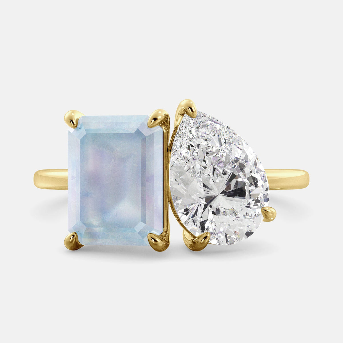 A toi et moi ring with an emerald-cut moonstone and a pear-cut gemstone. The emerald-cut morganite is set on the left of the ring, and the pear-cut gemstone is set on the right. The ring is made of recycled yellow gold and has a simple, elegant design. The pear-cut gemstone can be customized. **Here are some additional details about the image:** * The emerald-cut white moonstone is a birthstone for June. * The pear-cut gemstone can be customized to any stone of your choice.