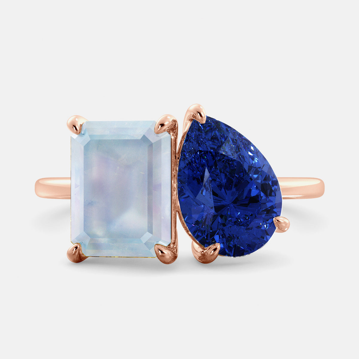 A gold ring featuring a rectangular blue moonstone on one side and a pear-shaped blue sapphire on the other, set in a yellow metal band.