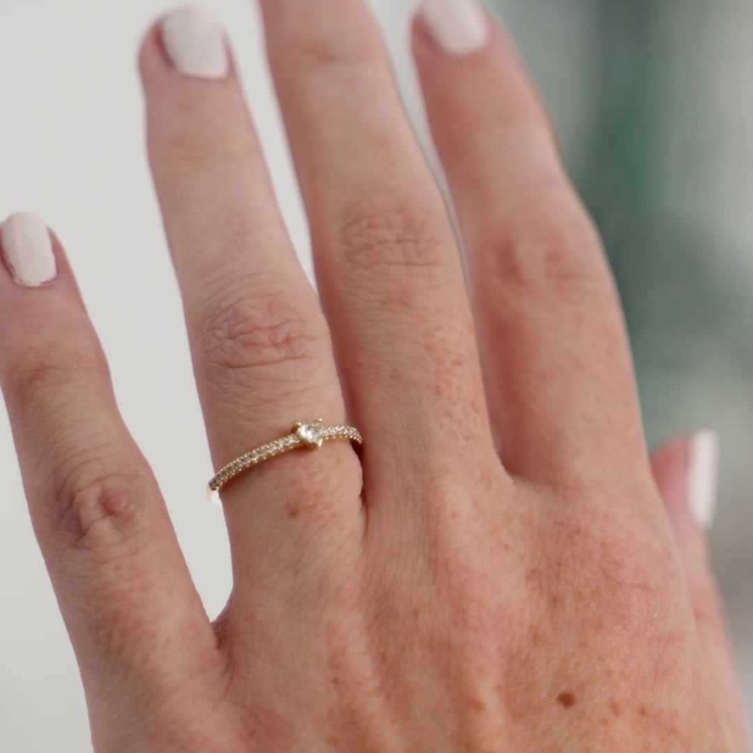 A close-up of a heart-shaped diamond ring with side stones in recycled yellow gold. The ring is set with a single, large diamond in the center of a heart-shaped setting. The ring is made of recycled yellow gold and has a simple, elegant design. The diamond is sparkling and reflects light beautifully. The side stones are smaller diamonds that are set in the band of the ring. The ring is a perfect gift for a loved one or a special occasion.