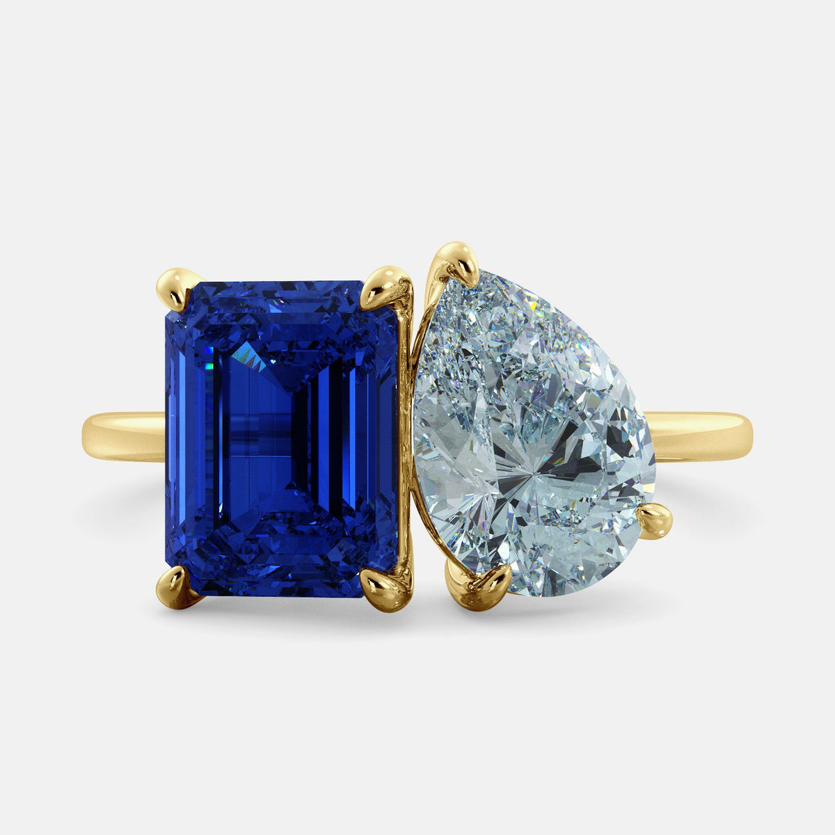 A toi et moi ring with an emerald-cut Blue Sapphire and pear-cut gemstone. The emerald-cut morganite is set on the left of the ring, and the pear-cut gemstone is set on the right. The ring is made of recycled yellow gold and has a simple, elegant design. The pear-cut gemstone can be customized. **Here are some additional details about the image:** * The emerald-cut white Blue Sapphire is a birthstone for September. * The pear-cut gemstone can be customized to any stone of your choice.