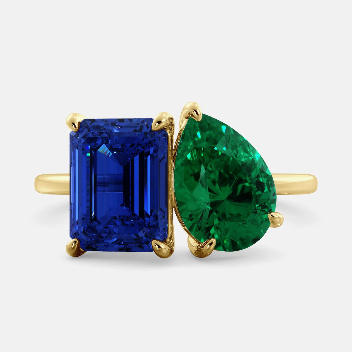 This image shows a toi et moi ring with a pear-shaped emerald and an emerald-cut blue sapphire. The ring is set in a 14k yellow gold band and is available in all sizes. The blue sapphire is a precious gemstone that is said to promote loyalty, wisdom, and truth. The emerald is a precious gemstone that is said to promote love, compassion, and forgiveness. The toi et moi ring is a beautiful and unique way to show your love and commitment to someone special.