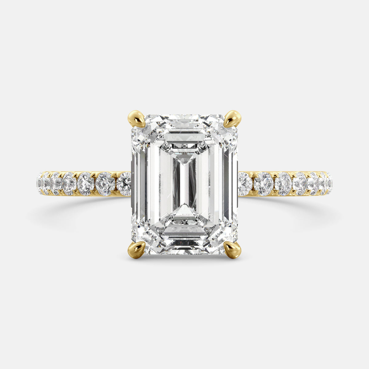 A close-up of an emerald solitaire lab-diamond ring with pave in yellow gold. The ring is set with an emerald-cut lab diamond in the center, surrounded by a row of smaller diamonds on the band. The diamond is climate-positive as it used carbon from the atmosphere to produce. The ring is available in a variety of sizes and starts at \\$6,350.00.