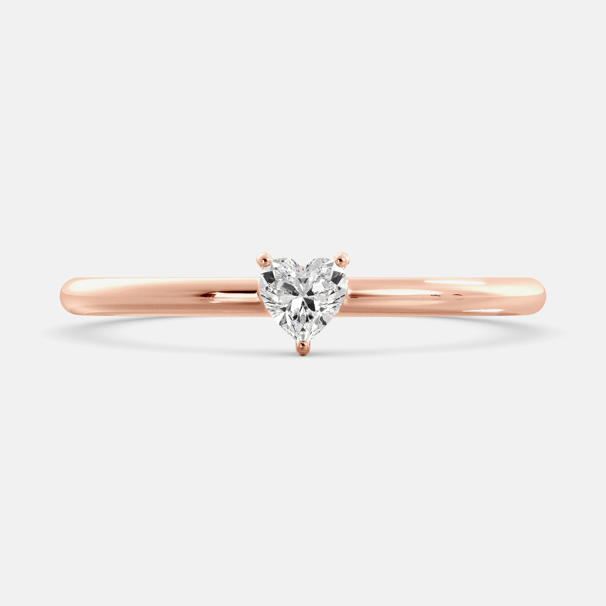 A close-up of a heart-shaped solitaire diamond ring in recycled rose gold. The ring is set with a single, large diamond in the center of a heart-shaped setting. The ring is made of recycled rose gold and has a simple, elegant design. The diamond is sparkling and reflects light beautifully. The ring is a perfect gift for a loved one or a special occasion.