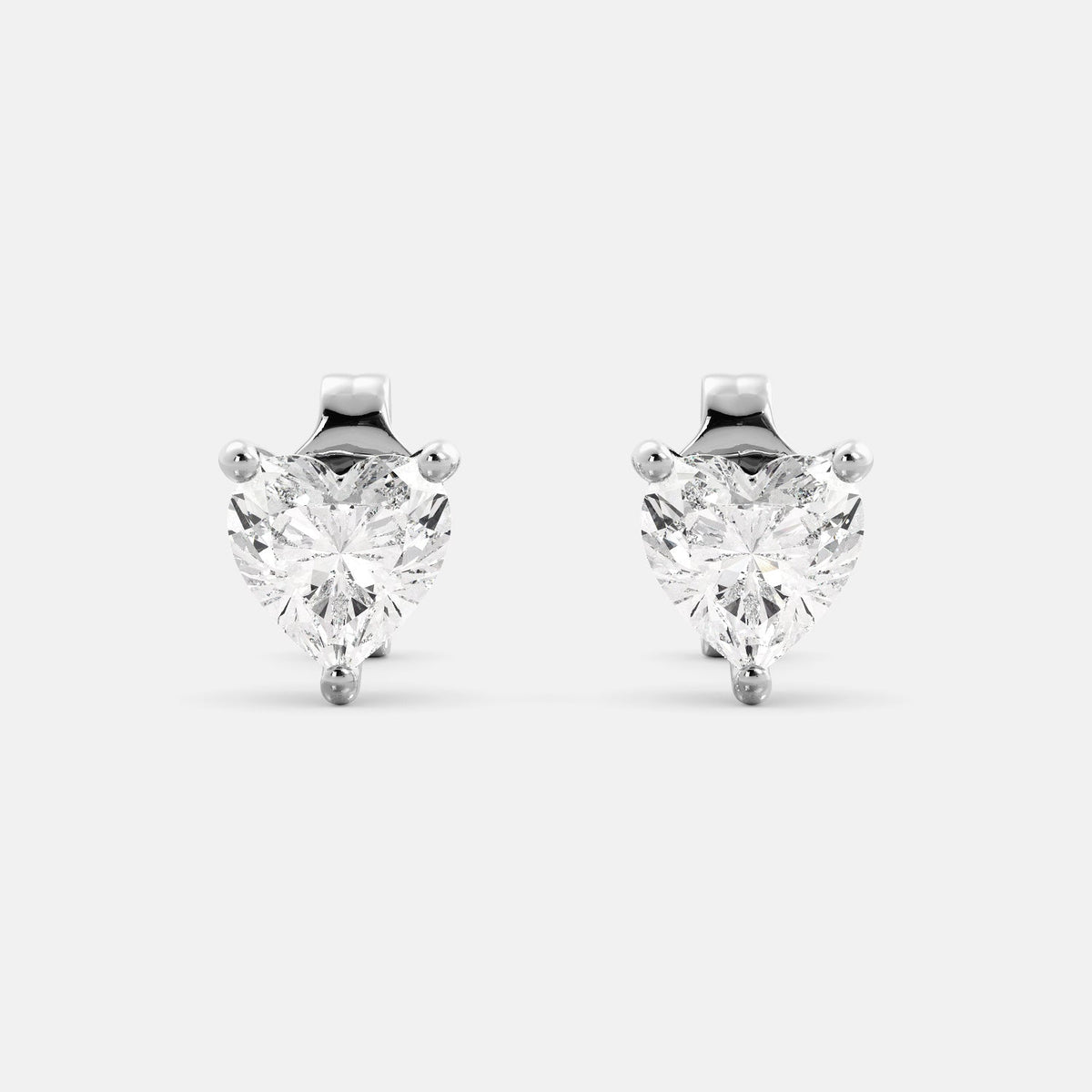 A close-up of a pair of heart-shaped diamond stud earrings in recycled white gold. Each earring is set with a single heart shape diamond in the center. The earrings are made of recycled white gold and have a simple, elegant design. The diamonds are sparkling and reflect light beautifully. The earrings are a perfect gift for a loved one or a special occasion.