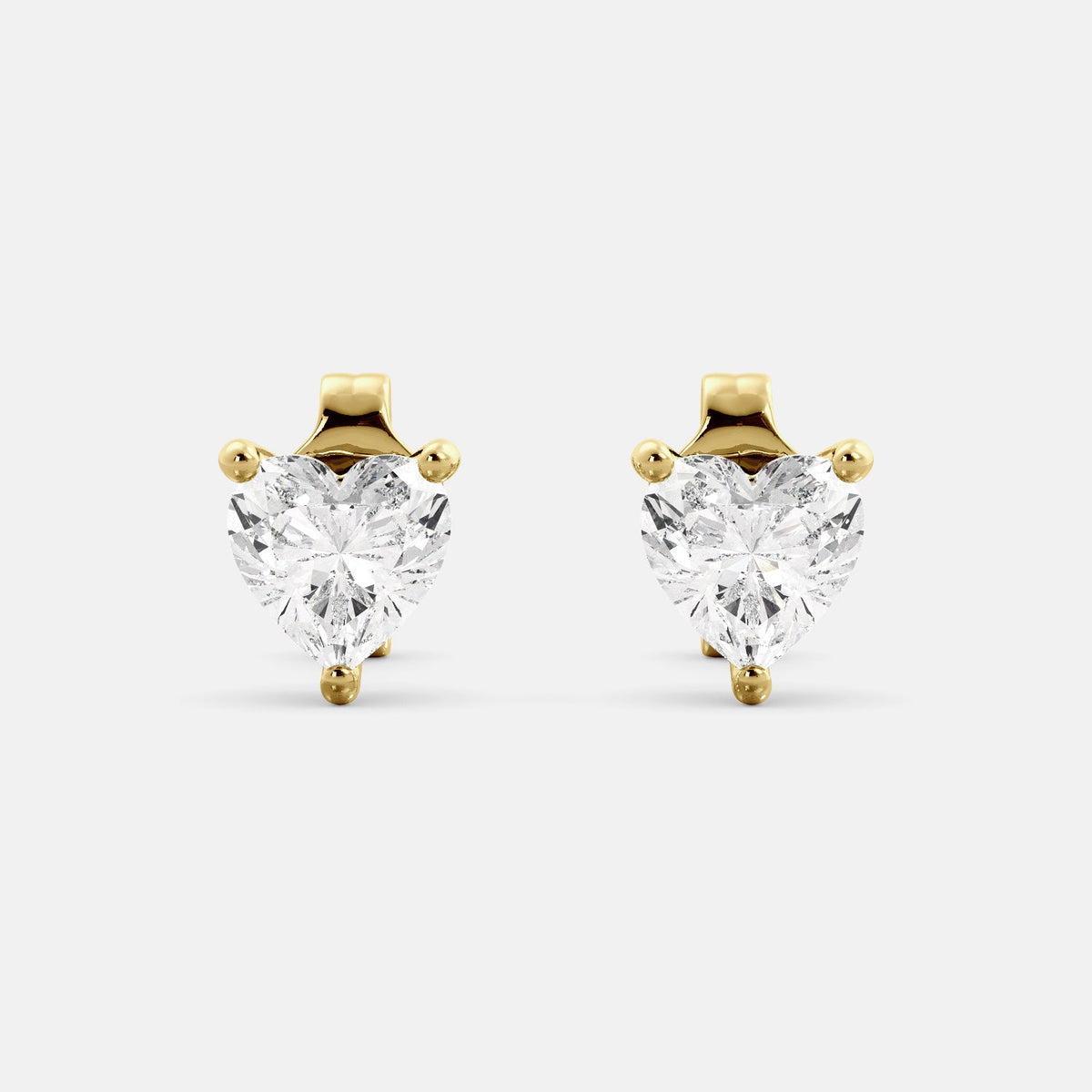 A close-up of a pair of heart-shaped diamond stud earrings in recycled yellow gold. Each earring is set with a single heart shape diamond in the center. The earrings are made of recycled yellow gold and have a simple, elegant design. The diamonds are sparkling and reflect light beautifully. The earrings are a perfect gift for a loved one or a special occasion.