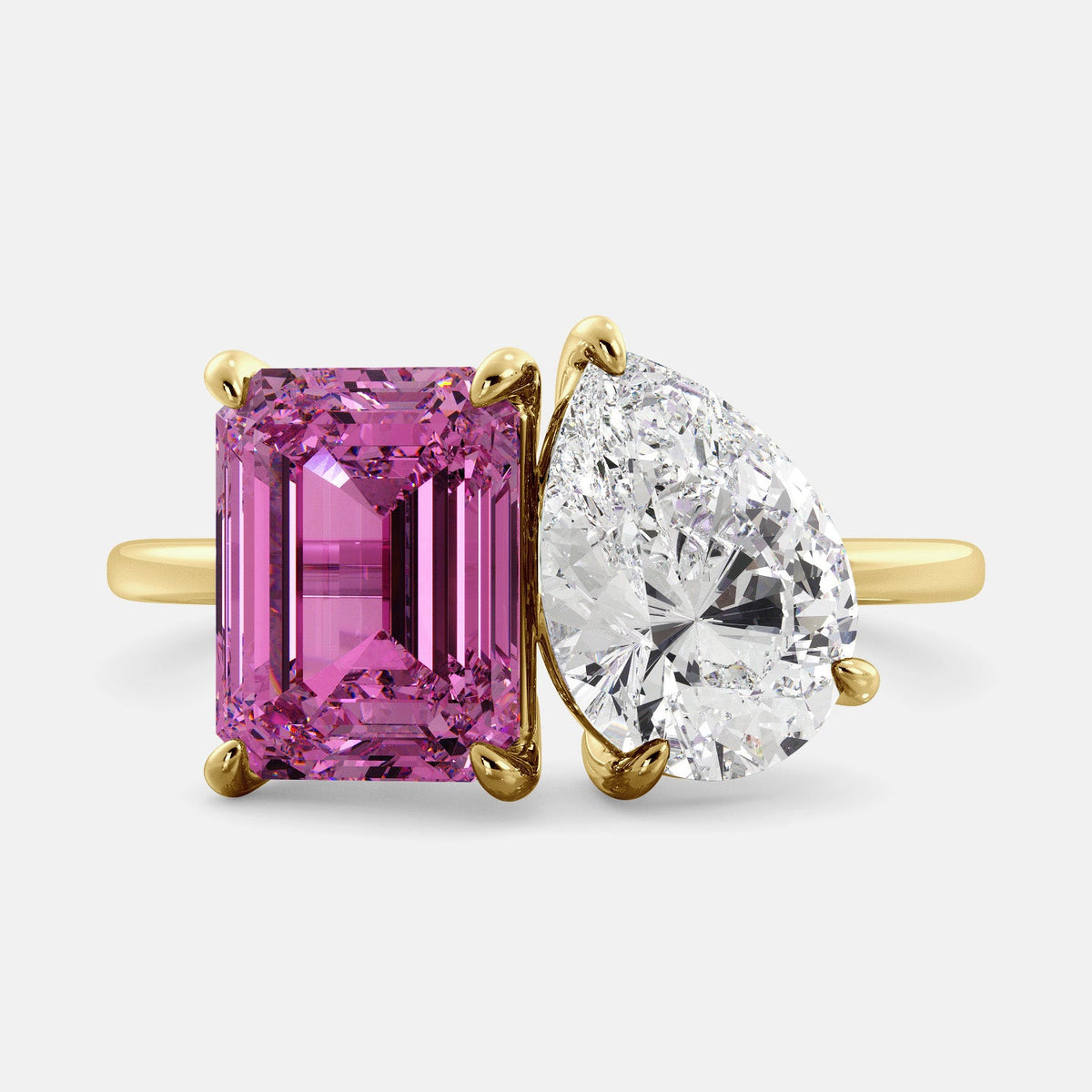 A toi et moi ring with an emerald-cut Pink Sapphire and pear-cut gemstone. The emerald-cut is set on the left of the ring, and the pear-cut gemstone is set on the right. The ring is made of recycled yellow gold and has a simple, elegant design. The pear-cut gemstone can be customized. **Here are some additional details about the image:** * The emerald-cut white Pink Sapphire is a birthstone for October. * The pear-cut gemstone can be customized to any stone of your choice.